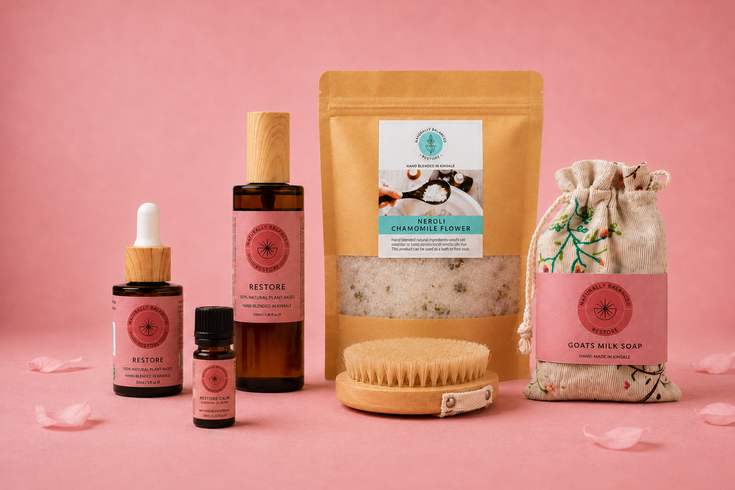 A curated skincare set arranged on a pink background, including amber dropper bottles, a large amber pouch with a visible bath salt window, a natural bristle brush, and a drawstring soap bag labeled 'Coated Milk Soap'. The products feature pink labels and wooden caps.