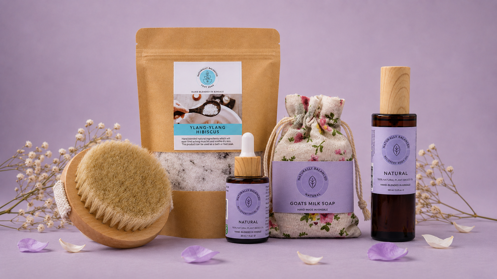 A spa gift set featuring a brown stand-up pouch, a small dropper bottle, a floral drawstring soap bag, and a dark amber bottle with a light wood cap, arranged with a wooden scrub brush and dried flowers on a lavender background.