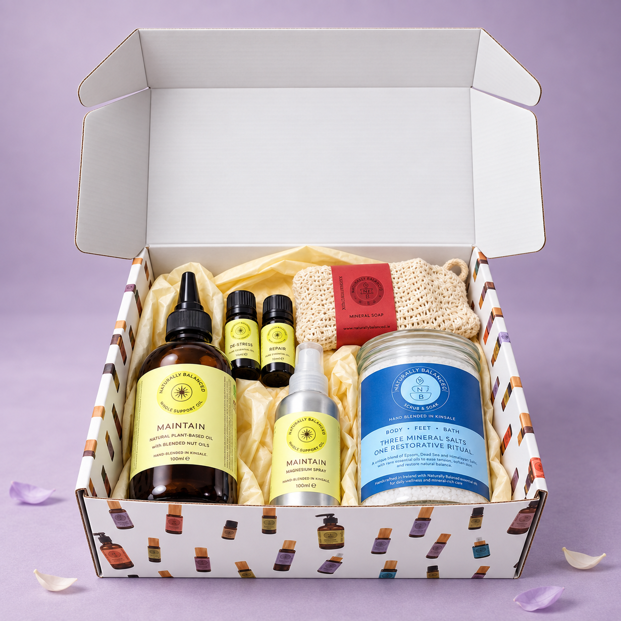 A curated skincare gift box from The Rituals, shown open against a lavender background. The box contains a brown glass bottle of hemp seed oil, three small essential oil dropper bottles, a silver mist spray, a blue tin of beeswax salt, a red and beige loofah, and a white patterned box lined with yellow tissue paper.