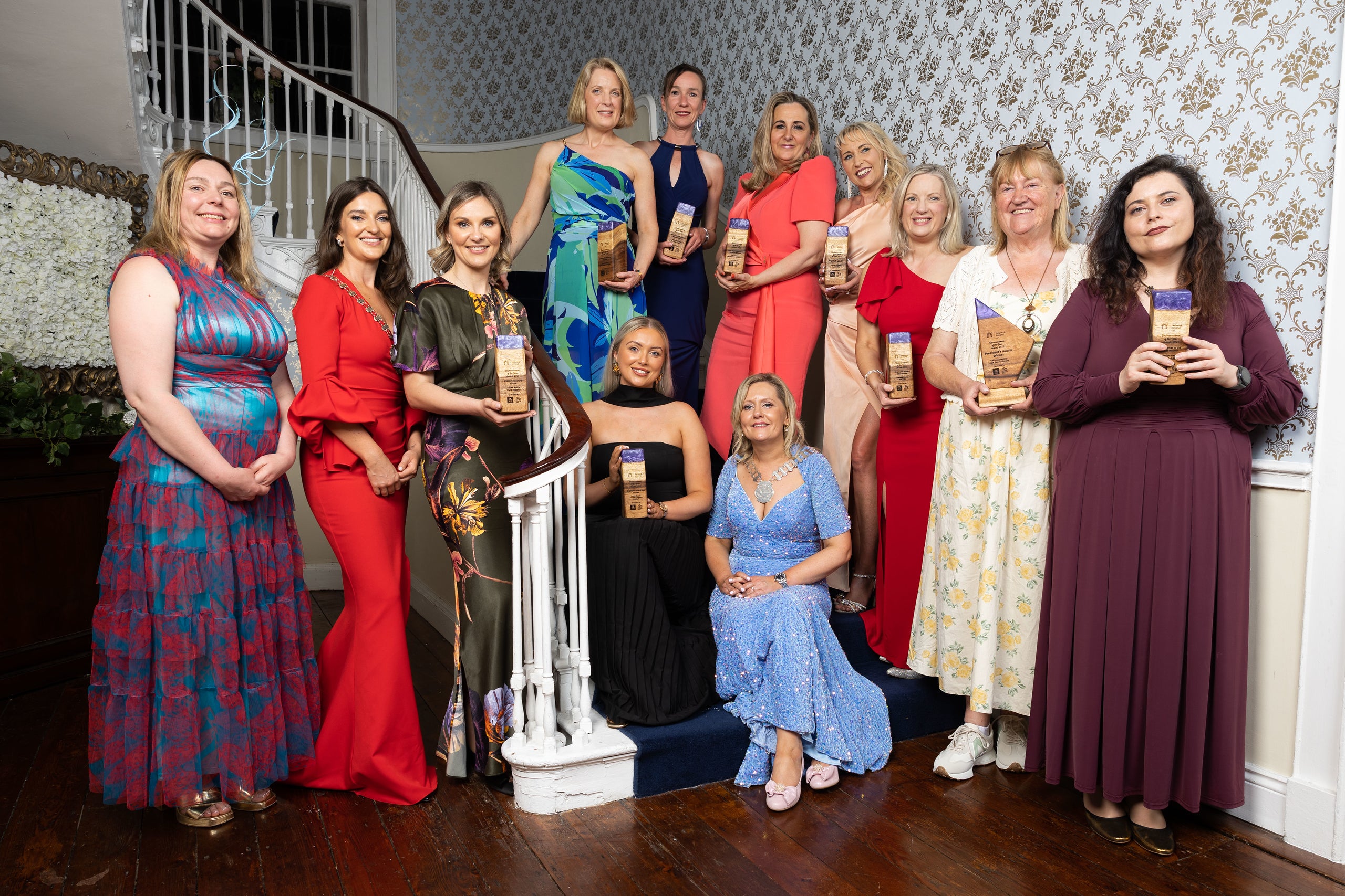 Group of women in formal attire standing on a staircase, holding awards.