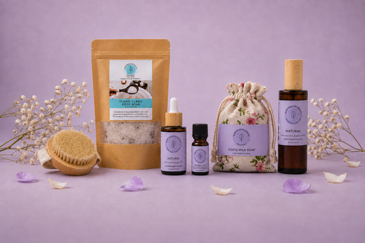 A spa gift set featuring a gold stand-up pouch, two amber dropper bottles, a floral drawstring bag, and an amber bottle with a wooden cap, arranged on a lavender surface with dried flowers and petals against a lavender background.
