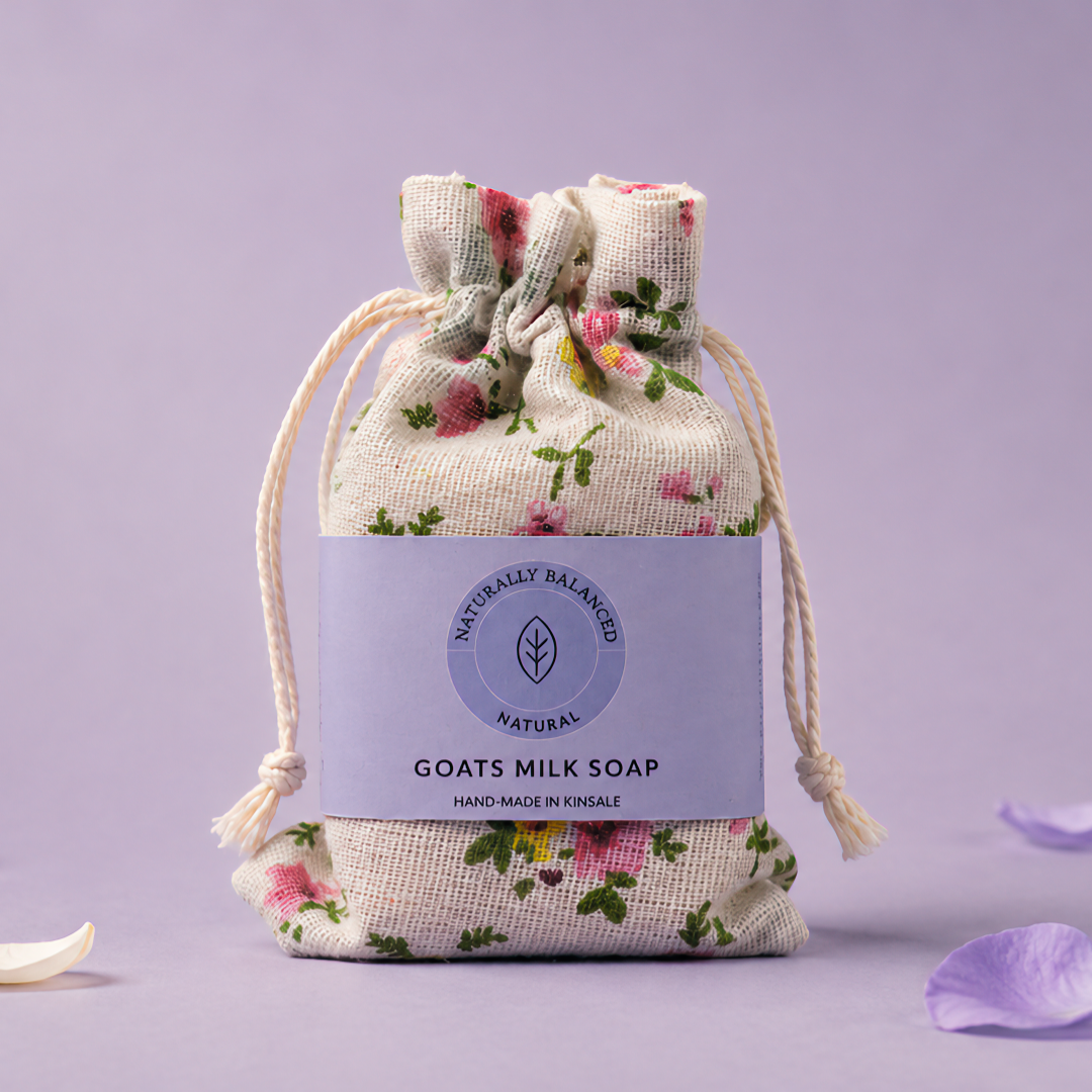 Goats milk soap in a floral drawstring bag on a light purple background with scattered petals.