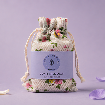 Goats milk soap in a floral drawstring bag on a light purple background with scattered petals.