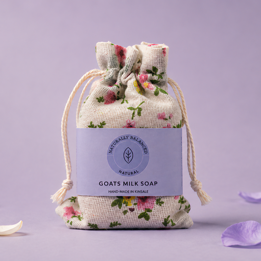 Goats milk soap in a floral drawstring bag on a light purple background with scattered petals.