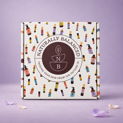 A square product box for Naturally Balanced skincare, featuring a cream-colored cover decorated with a colorful pattern of small cosmetic bottles and a large dark brown circular logo in the center. The box is photographed on a lavender surface with scattered flower petals.
